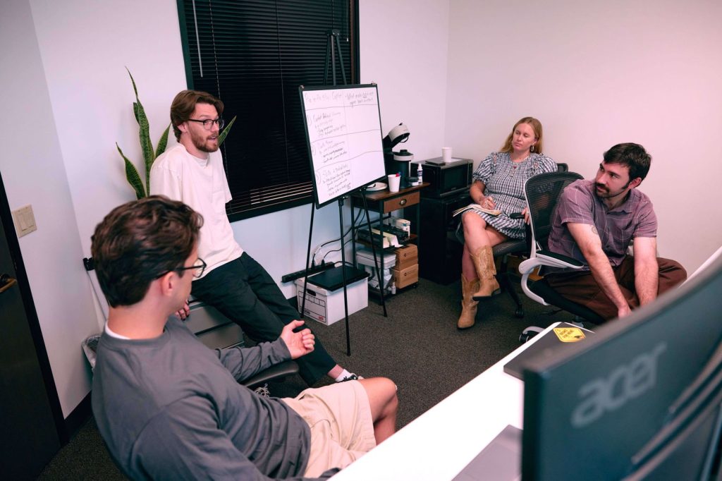 Four people having a discussion in a small office with a whiteboard and computer monitors.