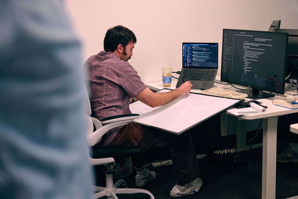 Man sitting at desk working on a laptop and a desktop monitor displaying code and a messaging app.