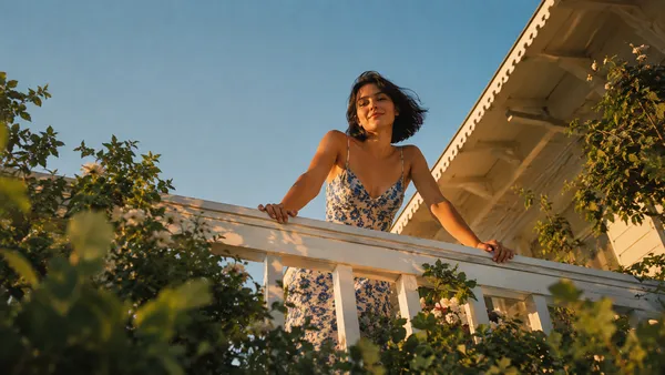 Wide low-angle shot looking up at young woman leaning over white wooden balcony railing of a charming house exterior, full figure visible from waist up, blue and white floral sundress, both hands on railing, short dark hair caught by breeze, lush green garden foliage framing the bottom and sides, bright blue sky filling upper background, warm golden afternoon light, negative space in sky for caption text, dreamy summer editorial photography, vertical 4:5 composition --ar 4:5 --v 6
Same extreme low-angle balcony peeking composition at golden hour, young woman with short dark hair peeking over white railing, warm amber-golden light washing over skin and floral dress, sky shifting from deep amber at horizon to blue above, blurred garden foliage in warm green-gold tones in foreground, long shadow cast across the railing, direct playful eye contact, cinematic golden hour color grade, vertical 4:5