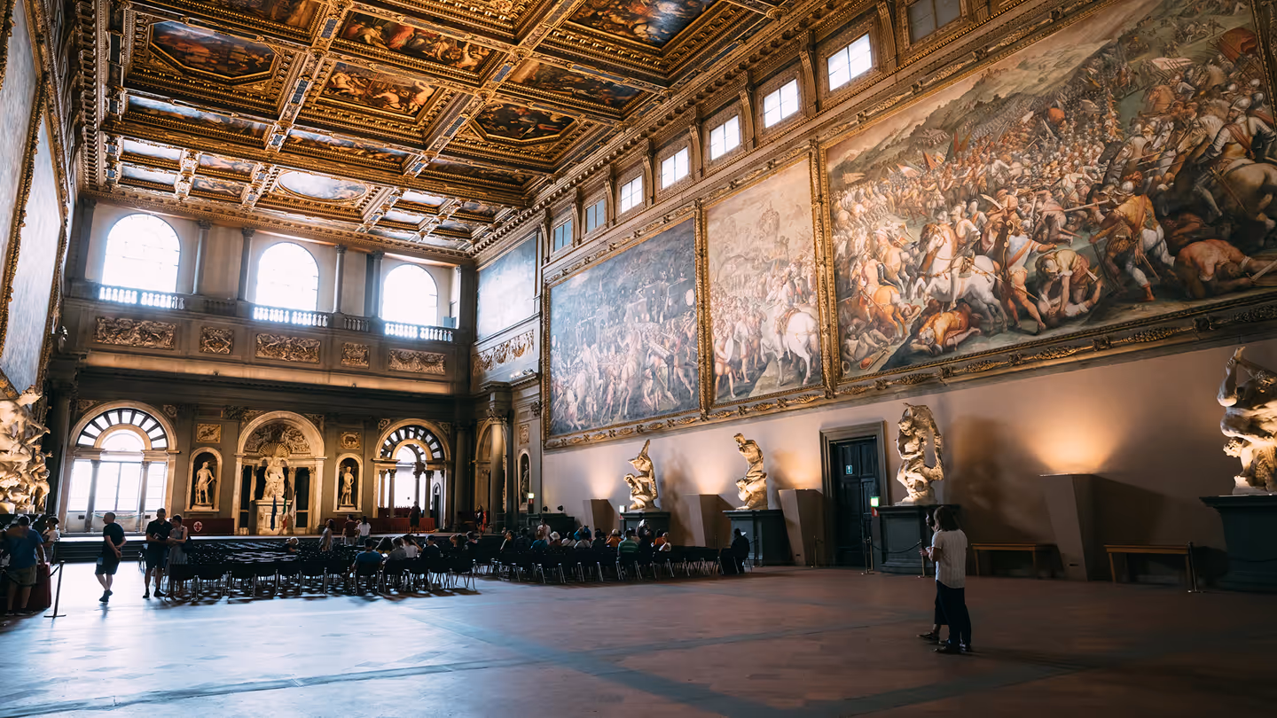Spacious museum hall with large historic battle murals, sculptures on pedestals, and visitors seated and walking.